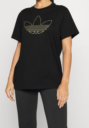 Woman wearing a black t-shirt with a gold Adidas trefoil logo and black pants against a plain light background.