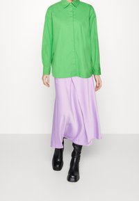 Grüne Hemdjacke mit langen Ärmeln, kombiniert mit einem fließenden lila Rock und schwarzen Knöchelstiefeln, vor einem schlichten weißen Hintergrund.