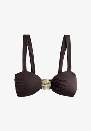 Kvinde i en mørk bikini går langs en solbelyst strand med vand, der reflekterer lys i baggrunden, og kigger ned.