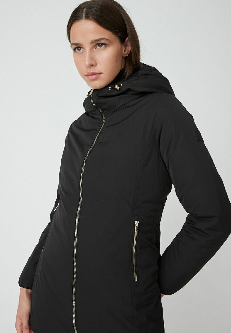 Zwarte gewatteerde jas met capuchon, voorzien van gouden ritsen, zijzakken en elastische manchetten. Gladde textuur, knielange snit.
