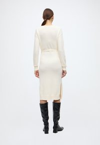 Πλεκτό φόρεμα εκρού με μακριά μανίκια, εφαρμοστή μέση, πλαϊνές σκισμές και ριμπ μανσέτες, συνδυασμένο με μαύρες μπότες έως το γόνατο.