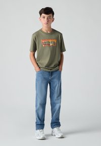 Camiseta de algodón verde oliva con un gráfico colorido de "Levi's", combinada con jeans de mezclilla azul claro. Zapatillas casuales completan el conjunto.