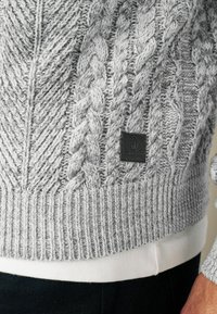Szary sweter z dzianiny z wzorem warkoczowym, ściągaczami na rękawach i teksturowanym korpusem. Zawiera czarną łatkę z logo w pobliżu brzegu.