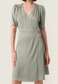Zavinovací šaty ze šedozelené jemně zvrásněné látky s krátkými nafouklými rukávy a výstřihem do V. Mají zavazování na boku a splývavou sukni.