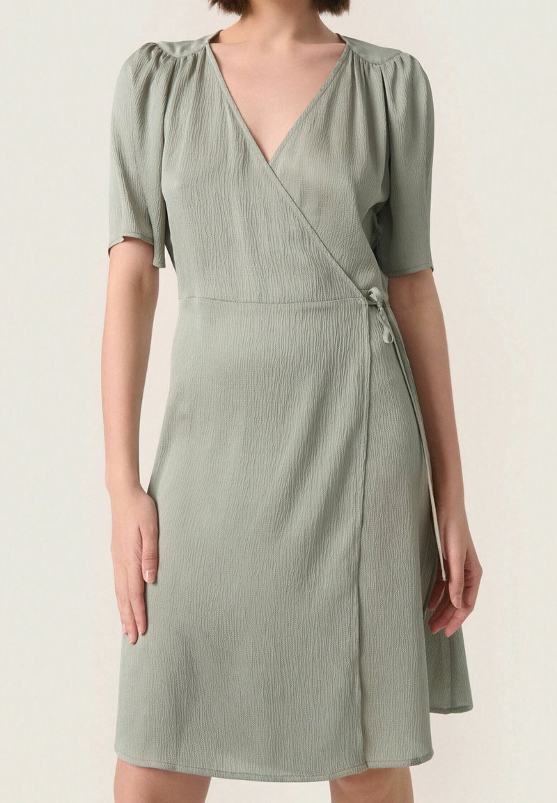 Zavinovací šaty ze šedozelené jemně zvrásněné látky s krátkými nafouklými rukávy a výstřihem do V. Mají zavazování na boku a splývavou sukni.