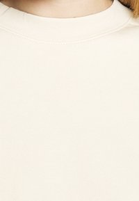 Sweatshirt beige chiaro con colletto a girocollo, texture morbida e dettagli di cucitura sottili, con una superficie liscia e uniforme.