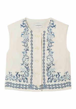Femme portant un haut crème sans manches avec broderie bleue, un pantalon crème taille haute avec ceinture chaîne dorée, et des mocassins crème sur fond blanc.