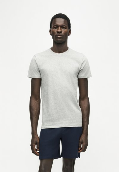 Un homme debout devant un fond blanc, portant un t-shirt gris clair à manches courtes et un short bleu marine, regardant droit devant avec une expression neutre.