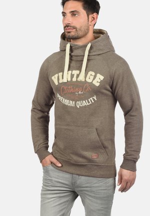 Sudadera marrón con cordones color crema, un bolsillo frontal y un diseño de texto de estilo vintage. La tela muestra una ligera textura; ajuste relajado.