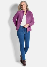 Lächelnde Frau mit grauem Haar, die eine lila Jacke, eine weiße Bluse, blaue Jeans und braune Stiefeletten mit Absatz trägt, im Vordergrund vor einem weißen Hintergrund steht.