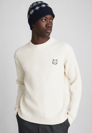 Man wearing white ribbed sweater with wolf logo and navy knit beanie, looking to the side with hands in dark trousers pockets.