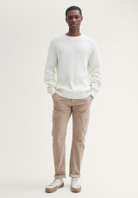 Chunky witte geribde trui, beige cargo broek met zijzakken en witte sneakers met beige accenten, staande tegen een neutrale achtergrond.