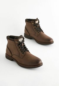 Brown leather boots feature a rounded toe, lace-up design, padded collar, and subtle stitching accents, mounted on a textured rubber sole.