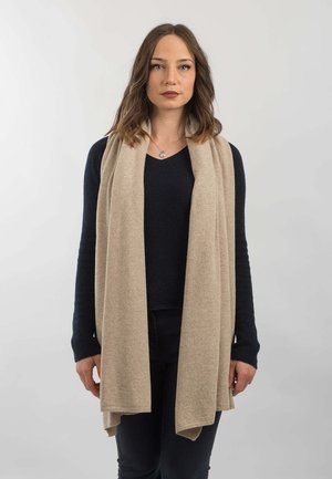 Beżowy dziany szalik, swobodnie ułożony na ciemnym swetrze, wyróżniający się miękką teksturą i smukłym, wydłużonym kształtem. Dodatki obejmują subtelny detal wykończenia.