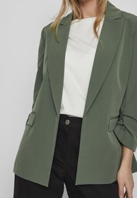 Zelený blazer vyrobený z lehkého materiálu, s klopovým límcem, ohrnovacími rukávy a postranními kapsami, v kombinaci s bílou košili a černými kalhotami.