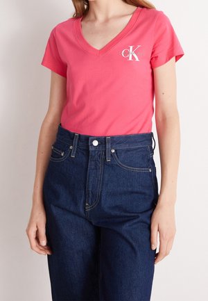 Kobieta w koralowo-różowej koszulce Calvin Klein Jeans z dekoltem w serek, włożonej do wysokich, ciemnoniebieskich jeansów na jednolitym tle.