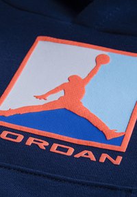 Marineblå sweatshirt med et struktureret Jordan-logo i orange og lyseblå, der fremviser en stiliseret spiller-silhuet og iøjnefaldende bogstaver.