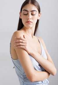 Giovane donna con lunghe capelli scuri, in un vestito senza maniche di un leggero blu, che tocca delicatamente la spalla sinistra su uno sfondo grigio uniforme.