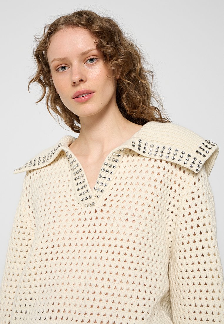 Cremfarbener Strickpullover mit V-Ausschnitt und großem Kragen, der mit metallischen Nieten entlang des Kragens verziert ist. Der Stoff hat eine offene Gewebestruktur.