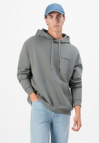 Grå hettegenser med kengurulomme og snorhette, med en svart logo. Kombinert med lyseblå jeans.