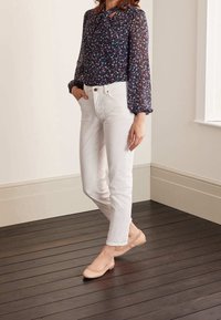 Blusa trasparente a maniche lunghe di colore blu navy con motivo floreale colorato, abbinata a pantaloni bianchi attillati e scarpe basse rosa chiaro su un pavimento in legno.
