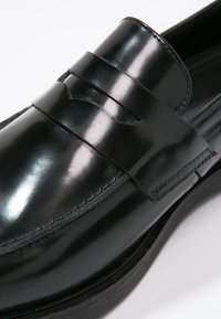 Mocassin en cuir noir avec une finition brillante, présentant une sangle décorative sur le cou-de-pied, des coutures discrètes et une semelle plate.