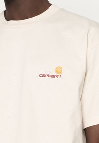 Beige cotton t-shirt featuring a crew neck and short sleeves. The front displays a small, orange-and-red embroidered Carhartt logo.