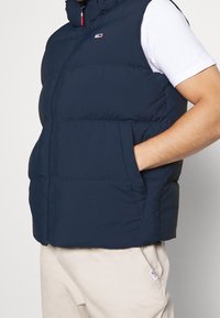 Granatowa puchowa kamizelka z wysokim kołnierzem i zapięciem na zamek; posiada boczne kieszenie i małe logo marki. Gładki, lekki materiał.