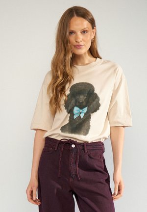 Frau trägt beige T-Shirt mit schwarzem Pudel und blauer Schleifen-Grafik, kombiniert mit hoch taillierter dunkelvioletter Hose mit Kordelzug in der Taille.