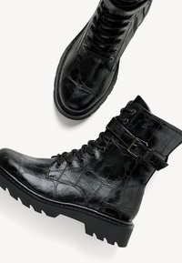 Bottes de combat en cuir noir avec un motif texturé, sangle à la cheville avec boucle, avant à lacets et semelle épaisse et robuste sur un fond blanc.
