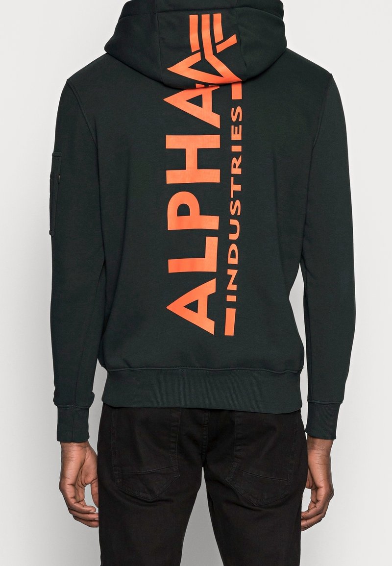 Sweat à capuche noir avec le texte "Alpha Industries" en orange vif disposé verticalement dans le dos, porté par une personne debout de dos.