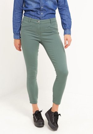 Dopasowane zielone spodnie z przednimi kieszeniami wsuwanymi, noszone z czarnymi sneakersami na platformie i niebieską dżinsową koszulą.