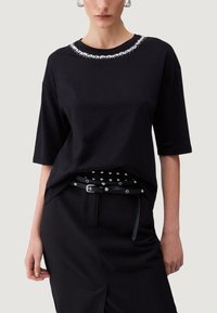 Nő fekete, rövid ujjú felsőt visel, melynek nyakánál ezüst díszítések találhatók, valamint fekete szoknyát, melyhez szegecselt öv tartozik a deréknál.