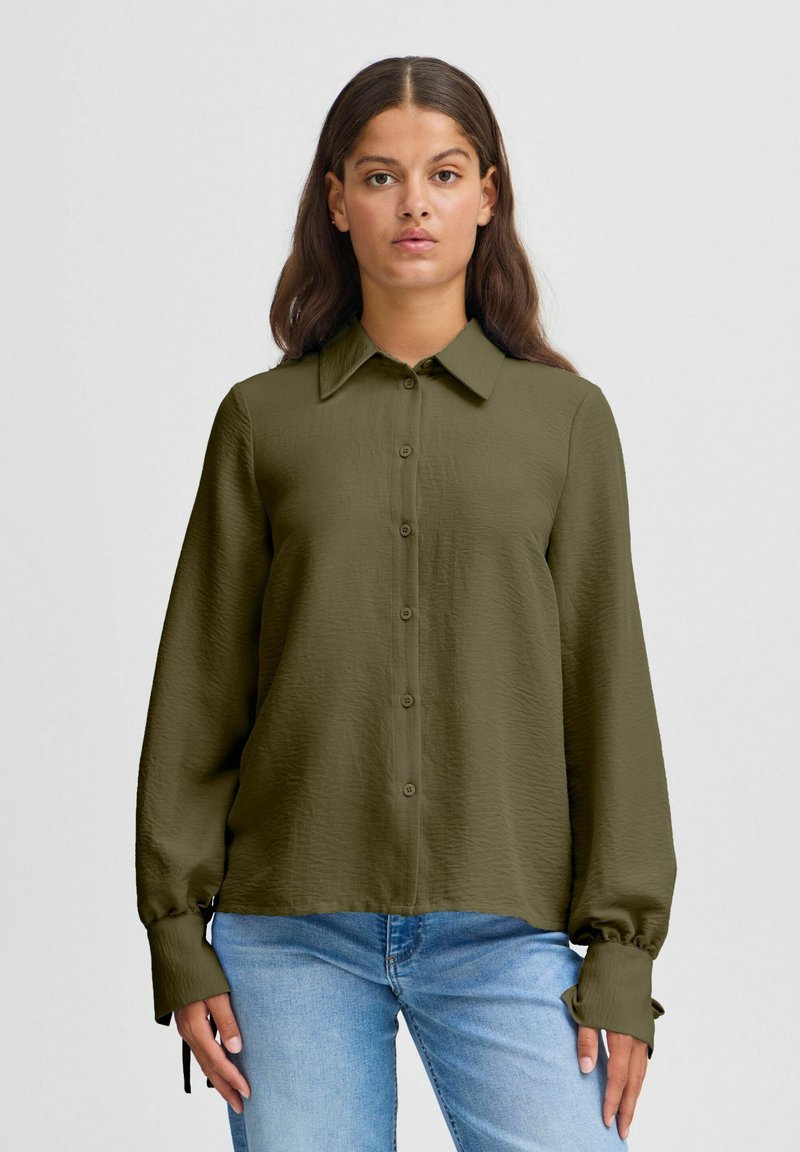 Olive green long-sleeve shirt with a button-up front, pointed collar, and gathered cuffs. Smooth, textured fabric. Paired with light blue jeans.