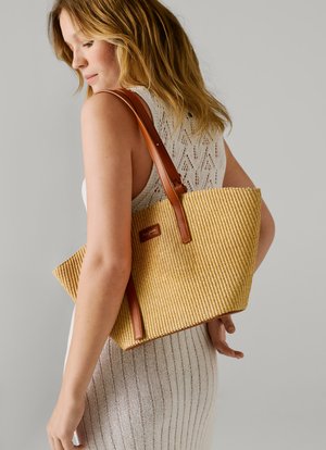 Žena sa plavom kosom nosi pletenu haljinu bez rukava i ima veliku tkanu torbu od slame s smeđim kožnim trakama preko ramena.