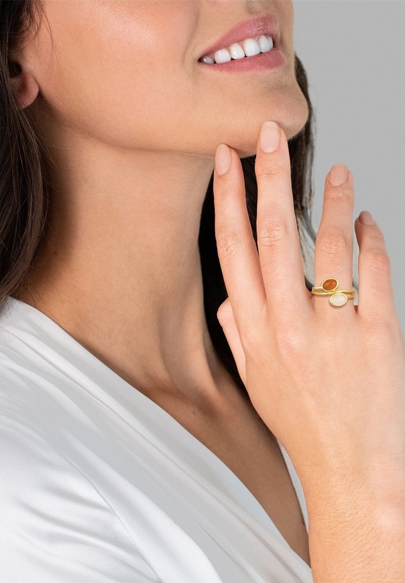 Goldfarbener Ring mit zwei Steinen: einem bernsteinfarbenen und einem cremigen. Schlankes Design mit glatter Textur, getragen an einer Hand vor einem hellen Hintergrund.