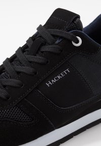 Baskets noires combinant du daim et du mesh, avec un col matelassé, des lacets ton sur ton, et une semelle extérieure blanche ornée du logo "HACKETT".