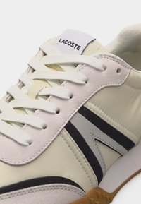 Közelkép egy bézs és fehér sportcipőről, fehér fűzőkkel, fekete és szürke oldalsávokkal, varrott részletekkel, valamint a nyelvén lévő "LACOSTE" felirattal.
