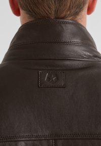 Braune Lederjacke mit hohem Kragen, ausgestattet mit einem gestickten Etikett auf der Rückseite und einer glatten, strukturierten Oberfläche.