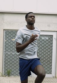 Grå kortærmet træningsskjorte kombineret med blå shorts. Løberen er i bevægelse foran en struktureret væg med et metalgittermønster.