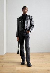 Schwarze Leder-Bomberjacke mit silbernen Details, hochgeschlossener schwarzer Oberteil, schwarze gerade geschnittene Jeans und schwarze Sneakers, hält eine kleine schwarze Tasche.