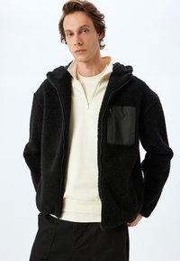 Chaqueta de forro polar negra con capucha, de tejido texturizado, y un bolsillo con cremallera en contraste en el lado izquierdo del pecho. Lleva una prenda de color beige claro debajo.