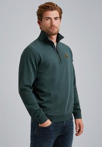 Dark green pullover sweatshirt with a half-zip collar, featuring a small logo patch, ribbed cuffs, and a smooth texture. Worn with jeans.
