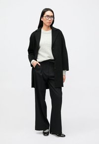 Čierny oversize kabát, sivý sveter a čierne nohavice s tenkými pásikmi. Model stojí sebavedomo, nosí okuliare a čierne topánky s hladkým povrchom.