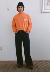 Oranžová košeľa s dlhými rukávmi, s predným vreckom a logom, v kombinácii s námorníckymi širokými nohavicami a hnedými topánkami, doplnená čiernou čiapkou.