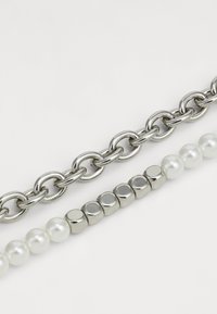 Des maillons de chaîne en argent au-dessus d'une rangée de perles brillantes et de disques métalliques, mettant en valeur un mélange de matériaux et de textures dans un design superposé.