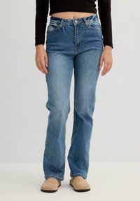 Vaqueros de mezclilla azules con cintura alta, corte recto y detalles en el dobladillo deshilachado. Llevados con una blusa negra de manga larga y zapatos beige.