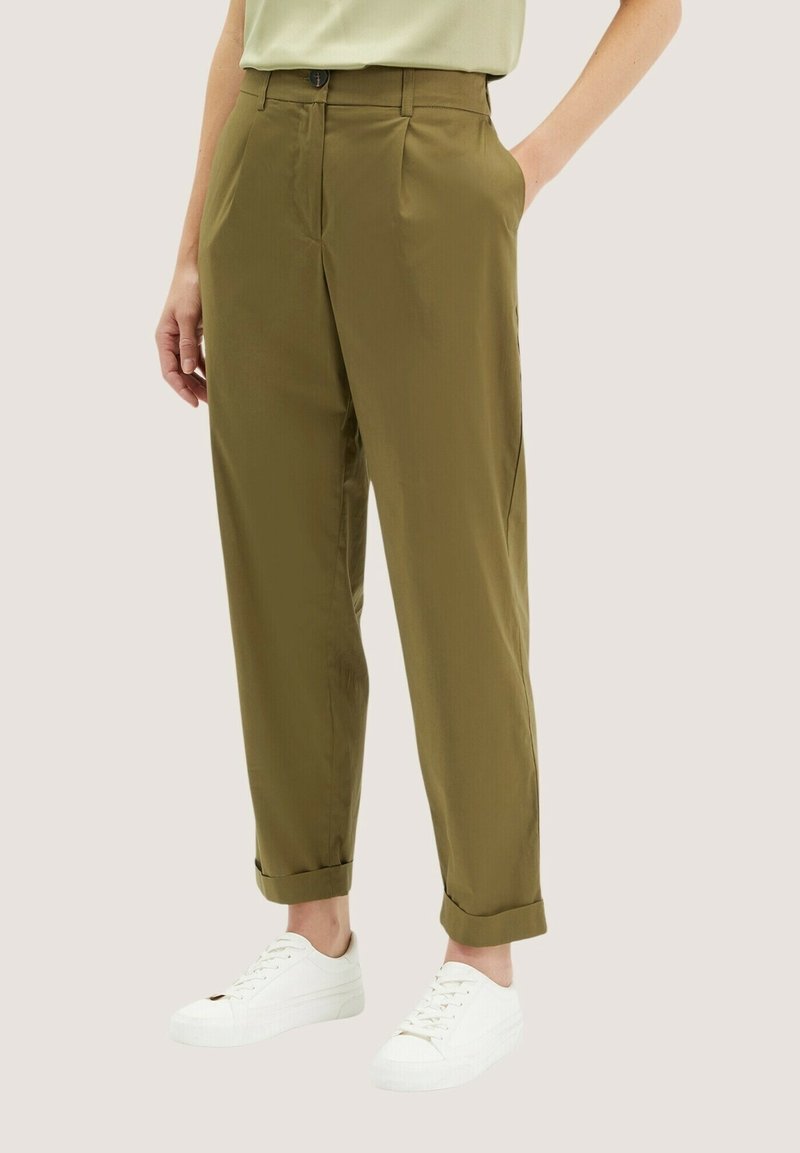 Personne portant un pantalon vert olive retroussé et des baskets blanches, debout avec une main dans la poche devant un fond uni.