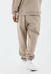 Joggers beige hechos de tejido suave presentan un corte relajado con puños ribeteados. Combinados con zapatillas blancas, mostrando detalles de cinturilla elástica.