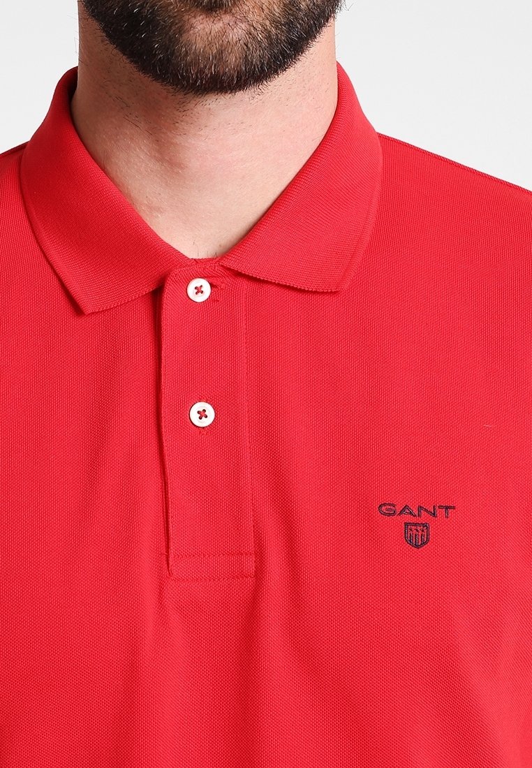 Rød polo skjorte med struktureret stof, tre-knaps lukning og kontrasterende hvide knapper. Har GANT-logo broderet i marineblå.